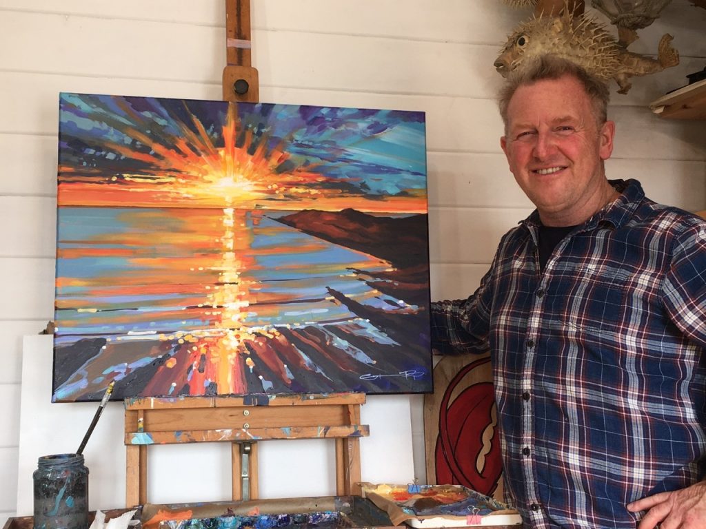 Woolacombe artist Steve PP standing at his easel in front a beautiful uplifting sunset painting.