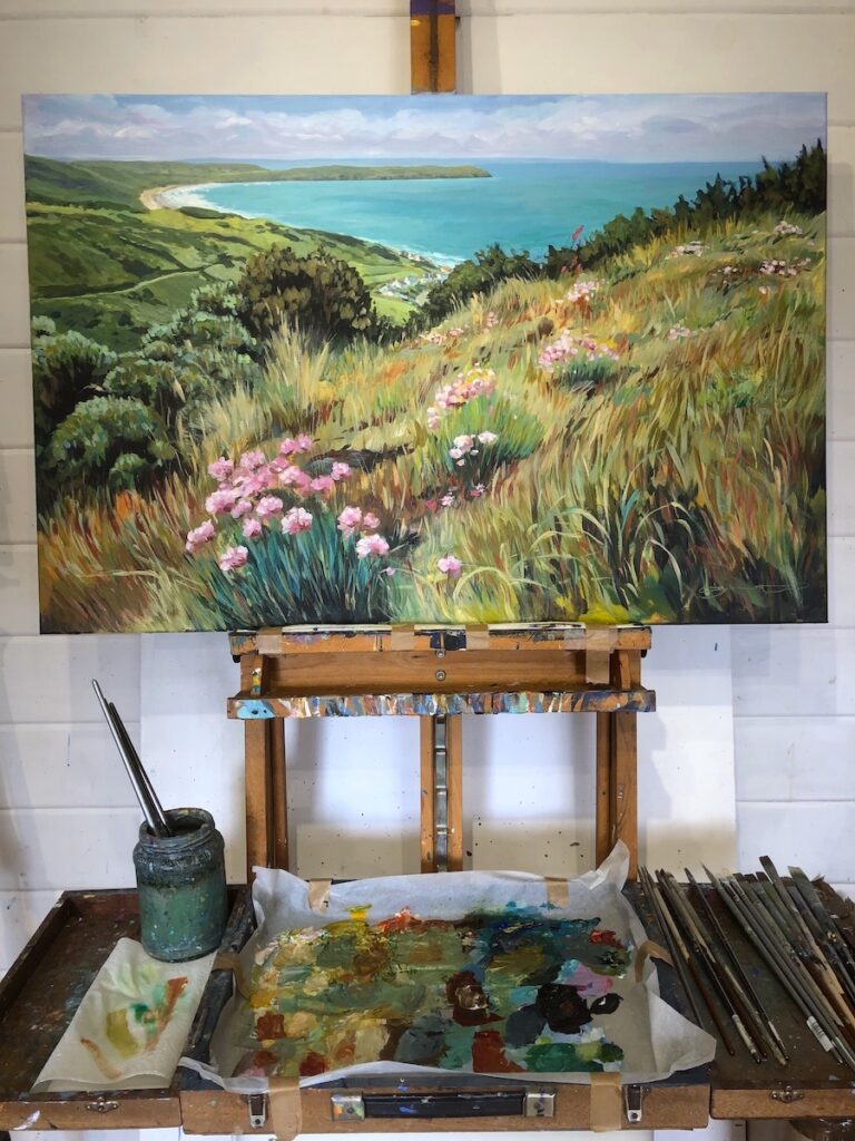 colourful painting of the view over the coast with sea pinks and gorse bushes in the foreground, on an artist's easel in a art studio